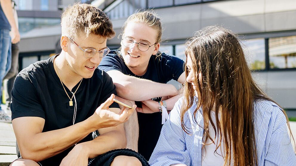 Zwei junge Männer und eine junge Frau schauen gemeinsam auf ein Smartphone